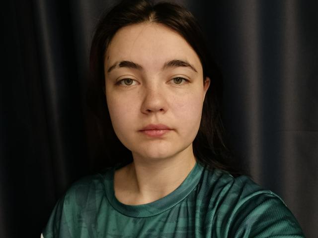 Marinda Howes hat helle Haut, grüne Augen und dunkelbraunes Haar. Sie trägt ein türkisfarbenes Hemd und blickt direkt in die Kamera mit einem neutralen Gesichtsausdruck. Ihre Brüste sind nicht sichtbar..