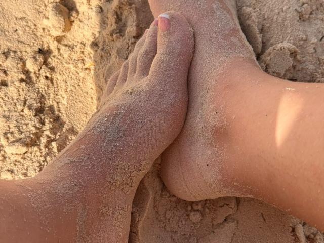 AdharaCooperr's sand-covered feet crossed on a sunlit beach, showing toes with pink nail polish, and her tan skin, with a slight shadow under her foot.