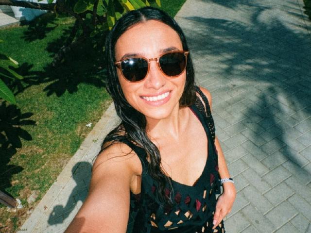 AdharaCooperr with tan skin, long wet black hair, brown aviator sunglasses, smiling, black lace top, visible cleavage, holding out phone for selfie, standing on gray brick path.