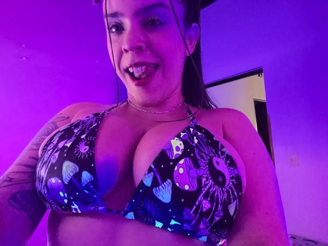 AliaPromax with large tits in a colorful, patterned bikini top, sticking out her tongue, purple-lit room, tattoo on right arm, long brown hair, and a subtle necklace.