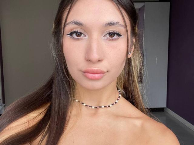 Brenda Bovia, con cabello castaño largo, ojos marrones y labios rosados, lleva un collar de cuentas, tiene un rostro lindo, y sus senos no son visibles en esta imagen..