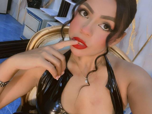 LindaFrancoo, avec des lèvres rouges, un haut noir brillant, un doigt dans la bouche, des yeux marron, des sourcils foncés, des cheveux ondulés, une chaise dorée, un collier, un tapis bleu, un piercing au nez, regardant la caméra..