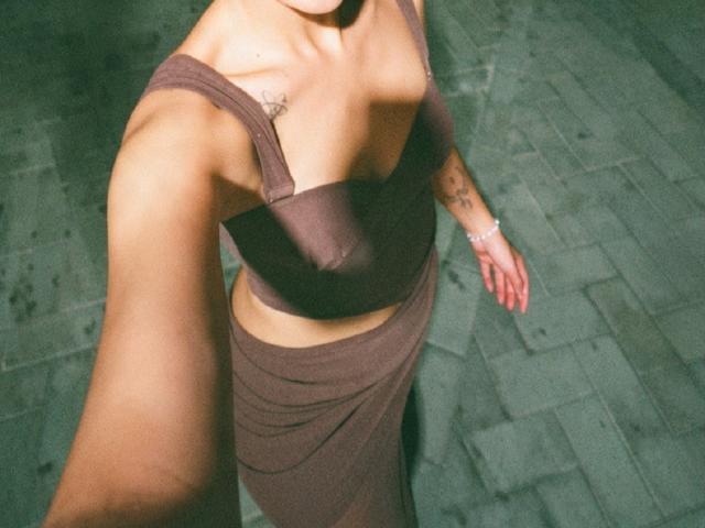 AdharaCooperr with bare tits in a brown top and matching skirt, holding a selfie, tattoo on right arm, bracelet on left wrist, green tiled floor, arms raised.