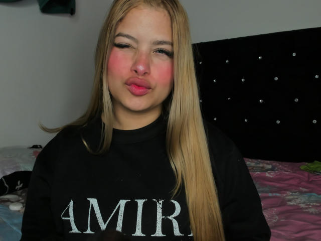 ShantalSummer has blonde hair, pink cheeks, and is wearing a black AMIR shirt. She's pouting with pink lips, sitting on a bed with a pink and floral bedspread.
