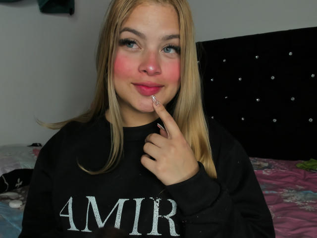ShantalSummer with blonde hair, pink cheeks, and black "AMIR" sweatshirt, touching her pink-lipped mouth with a finger, smiling slightly.