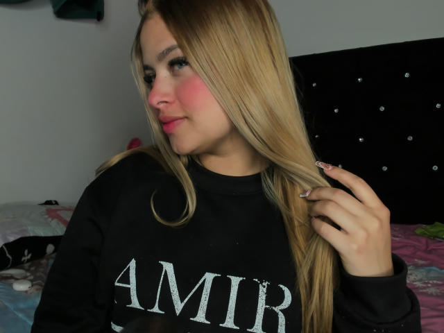 ShantalSummer with long blonde hair, pink blush, black AMIR sweatshirt, hand in hair, slight smile, pink lipstick, dark headboard, bed with pink and green blankets.