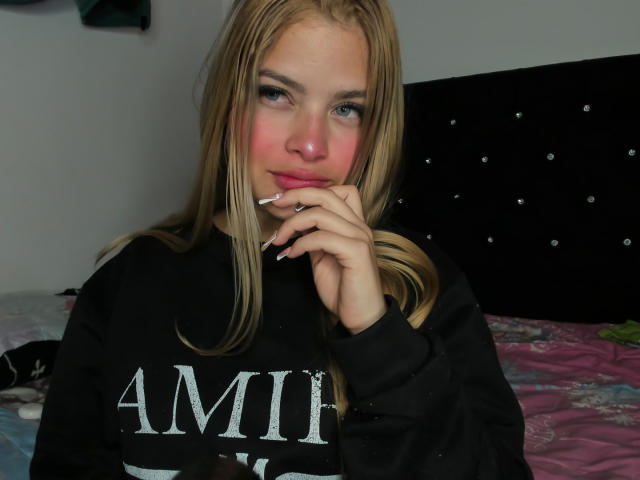 ShantalSummer with blonde hair, pink cheeks, and a cigarette in her mouth, wearing a black "AMF" sweatshirt, looking at the camera with a slight smile.