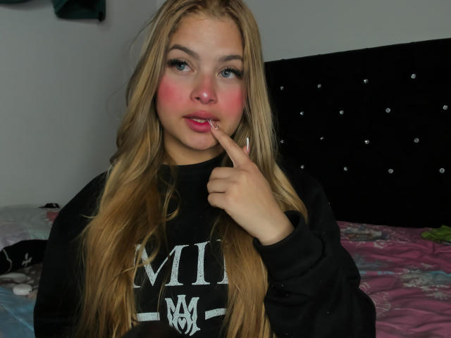 ShantalSummer with long blonde hair, pink blush on cheeks, black sweatshirt, finger touching pink lips, looking at camera, sitting on bed with pink and blue patterned sheets.