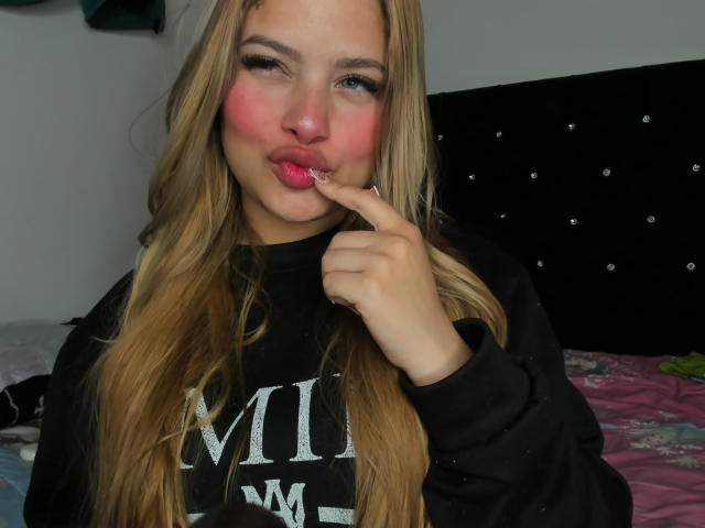 ShantalSummer with long blonde hair, puffy pink cheeks, black sweatshirt, and finger on lips, looking cute and playful, showing off her pink lipstick.