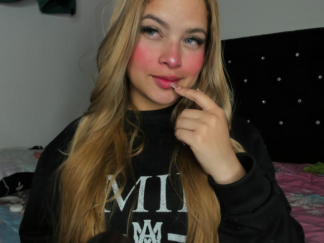 ShantalSummer with long blonde hair, pink blush on cheeks, black sweatshirt, and finger near pink lips, looking at camera, in bedroom with black headboard.
