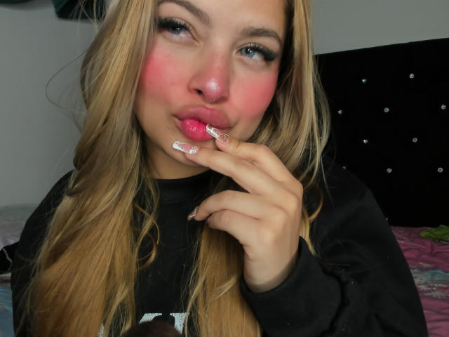 ShantalSummer with pink blush on her cheeks, pouty pink lips, blonde hair, black long-sleeve shirt, and white manicured nails, touching her lip.
