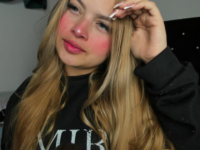 ShantalSummer with pink blush on her cheeks, long blonde hair, black shirt, pink lips, and white nails, slightly smiling, touching her head with her right hand.