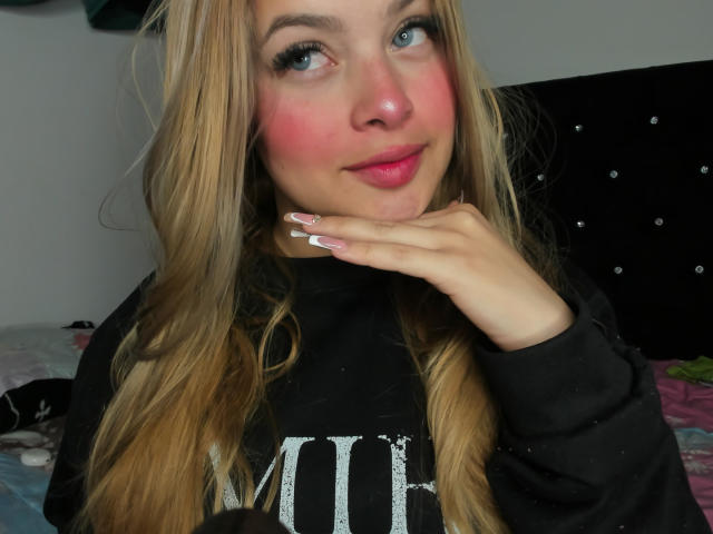 ShantalSummer has blonde hair, blue eyes, and pink cheeks, wearing a black hoodie, with her hand touching her chin, showing off her long, white-painted nails.