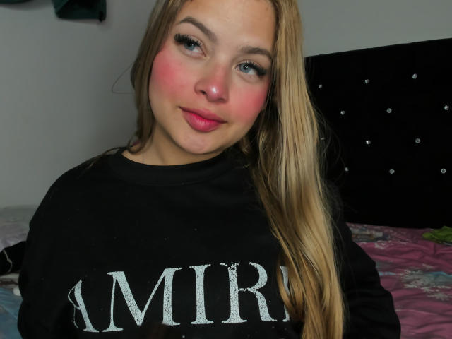 ShantalSummer has pink blushed cheeks, blue eyes, and long blonde hair. She's wearing a black sweatshirt, and her small tits are slightly visible. She's giving a cute, pouty smile.