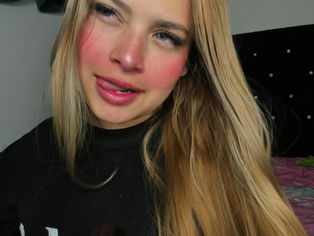 ShantalSummer has blonde hair, pink blush on her cheeks, and is sticking her tongue out with pink lips. She's wearing a black shirt, looking cute and slightly goofy.