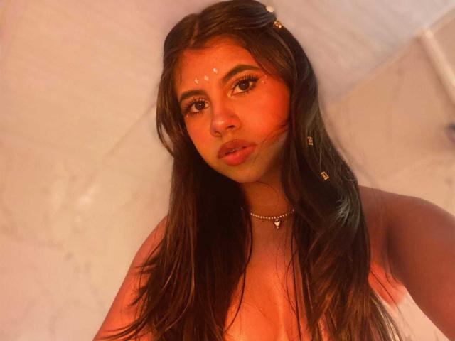 CelesteMorel has long brown hair, orange-tinted skin, and a face painted with white dots. She's topless with visible nipples, wearing a necklace, and has full, pouty lips.