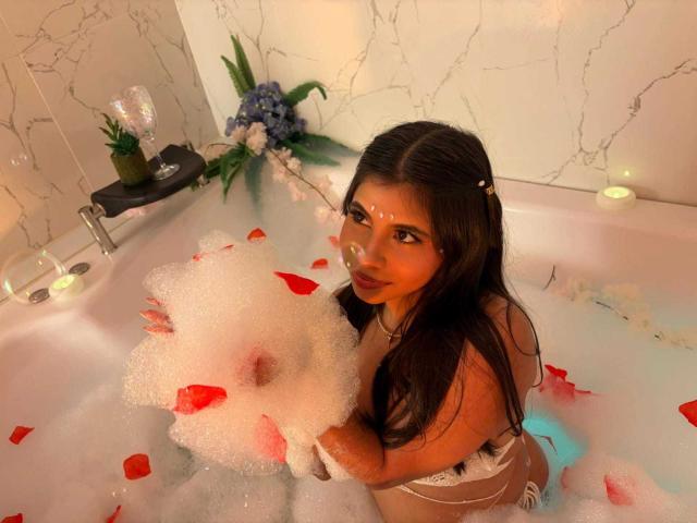 CelesteMorel with dark hair, glowing skin, and red dot on forehead, in a bubbly bathtub with rose petals, holding foam, looking at camera, candlelight in background.