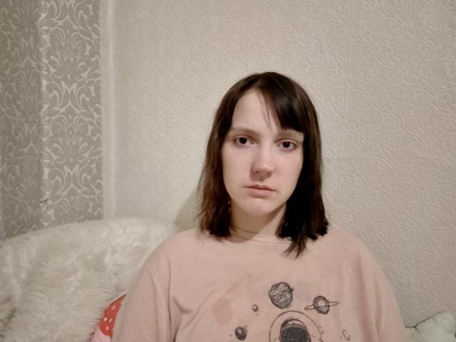VeldaMilbourn, con i capelli castano corti, pelle chiara, indossa una maglietta rosa con un motivo a tema spaziale, guarda direttamente la fotocamera, seduta su un cuscino bianco e soffice..