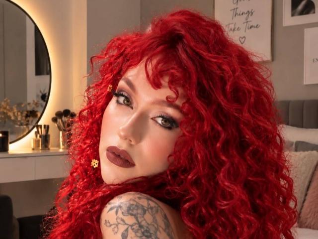 GaiaDoll with bright red curly hair, dark lipstick, and a tattoo on her right arm, looking back with a sultry expression. Her green eyes stand out.
