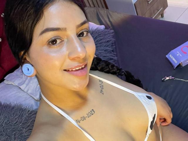 LonxEva with dark hair, smiling, wearing a white bikini top, showing small tits, tattoo on chest, blue ear gauges, lying on a bed with gray pillows.