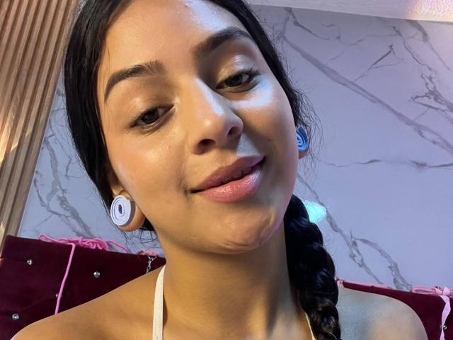LonxEva's face with smooth brown skin, dark eyes, and a slight smile. She's got earbuds in, a white halter top, and a braided black ponytail. Her lips are glossy.