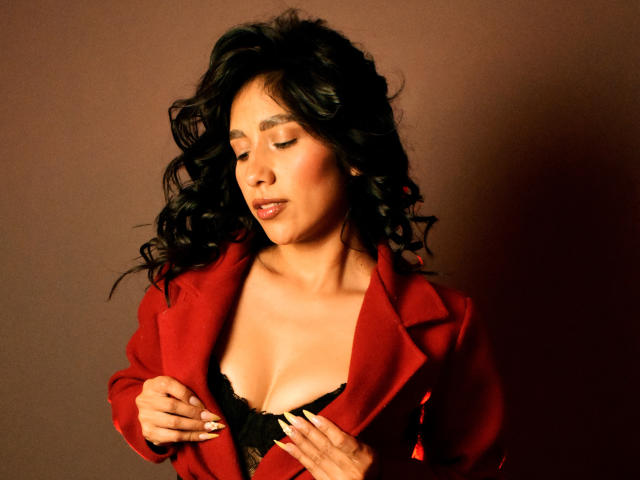 Briana, cheveux noirs bouclés, portant un blazer rouge, dévoilant un soutien-gorge en dentelle noire, les yeux fermés, les mains près de la poitrine, peau bronzée, sexy, confiante, fond marron, lumière douce..