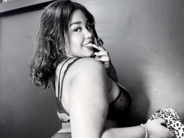 NicolZurich has curly dark hair, a smile, and is biting her finger. She's wearing a black bra, showing her big tits, and holding a leopard print cloth.