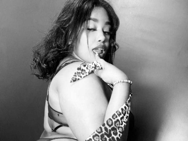NicolZurich with curly hair, tongue out, touching leopard-printed arm, wearing a leopard-printed bra, looking over shoulder, gray background, slightly seductive, wearing a pearl bracelet.