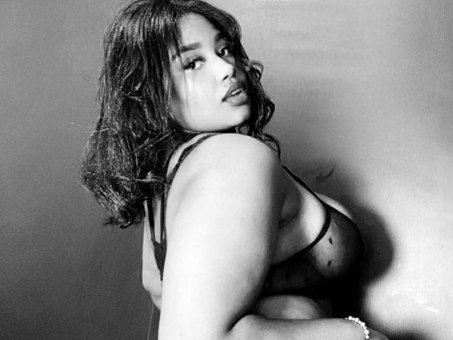 Black-and-white photo of NicolZurich with wavy hair, looking over shoulder, wearing a sheer black bra, showing her big tits, and a bracelet on her right wrist.