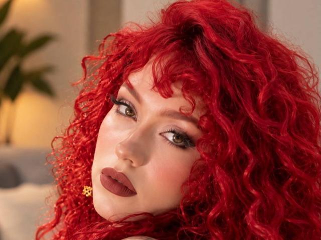 GaiaDoll with bright red curly hair, pale skin, green eyes, and dark red lipstick, looking seductively at the camera. Her nipples are hidden, but she's wearing a gold necklace.