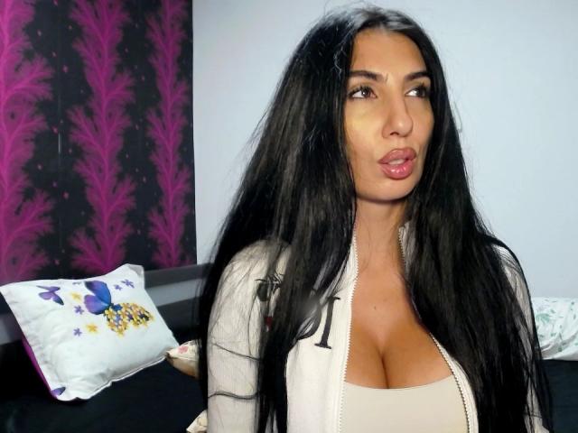 BrunetteBabe69 with long black hair, pouty lips, and big tits, wearing a white jacket over a beige top, looking to the side. She has a tan skin tone.