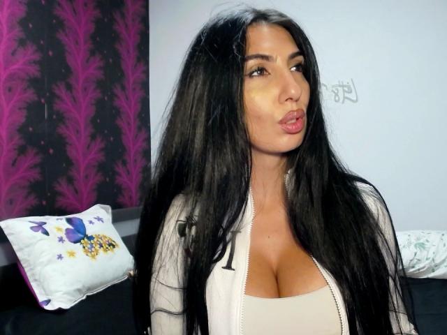 BrunetteBabe69, avec ses longs cheveux noirs, ses lèvres pulpeuses et ses grandes seins, porte un haut beige à décolleté plongeant et une veste blanche zippée. Elle regarde sur le côté et fait la moue..