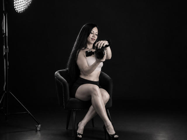 AmaraMirage with long black hair, wearing a white top, black shorts, and high heels, sits on a chair, holding a camera, looking at it, dark background, studio light.