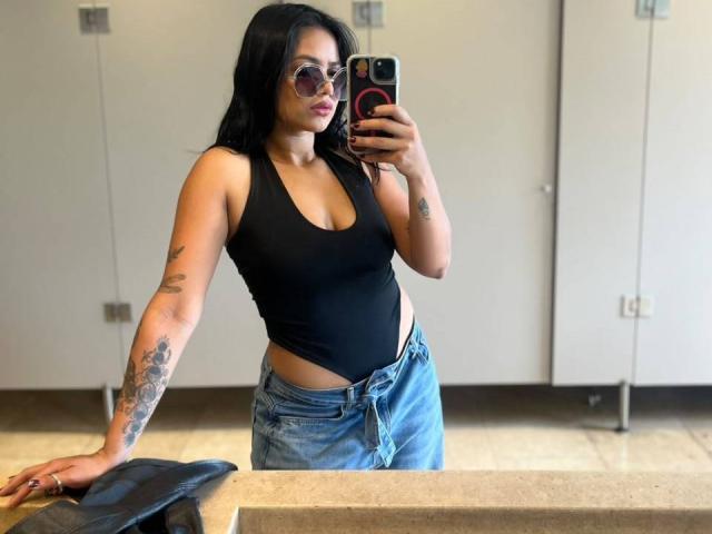 AlanaLuxe with black tank top, showing her tits in a tight fit, wearing blue jeans, tattoos on her arm, taking a mirror selfie, holding a phone with a pink case.