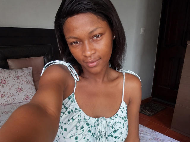 JuicyLeeX, con piel marrón media, cabello negro y liso, y senos pequeños, vestida con un vestido floral verde, tomando un selfie en un dormitorio, sonriendo suavemente, con el brazo extendido..