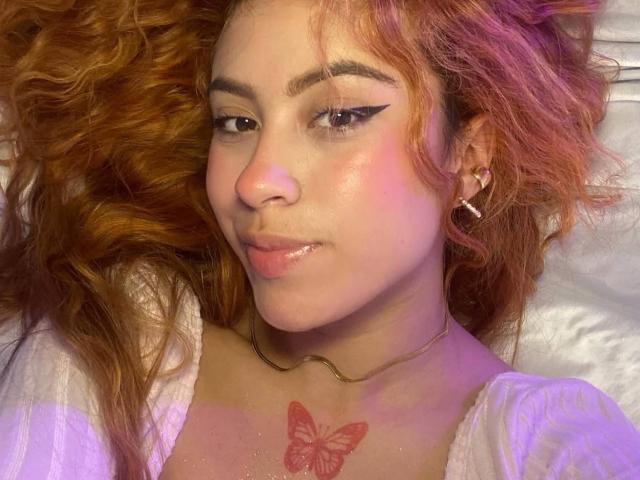 OriRivass with wavy orange hair, black eyeliner, pink blush, small nose, shiny lips, white shirt, gold necklace, butterfly tattoo on chest, earrings, smiling, laying down.
