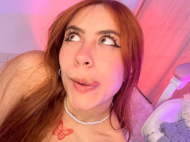 OriRivass has red hair, a pearl necklace, and a red butterfly tattoo on her chest. She's sticking out her tongue with wide eyes and long eyelashes.