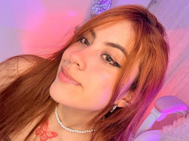 OriRivass with long red hair, light skin, and green eyes, wearing a pearl necklace and silver hoop earrings, smiling at the camera, showing a butterfly tattoo on her chest.