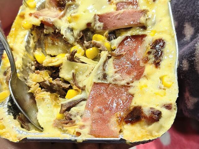 A cheesy casserole with crispy bacon, corn, and shredded meat, held in a metal tray with a spoon, looking super damn delicious. StephanieSun's hands are visible.