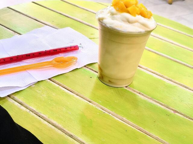 A green wooden table with a yellow drink in a glass, topped with orange pineapple chunks, red straw, and orange plastic spoon on a white napkin.