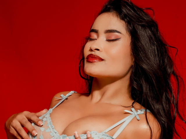 KarolPalacios with her eyes closed, wearing a light blue lace bra, touching her large tits, red lipstick, and wavy black hair, against a bright red background.