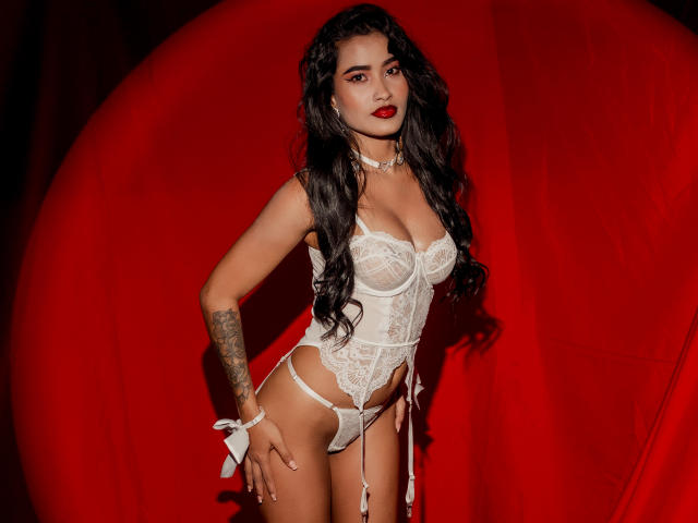 KarolPalacios in white lace lingerie, standing with hands on hips, long curly black hair, red lipstick, tattoo on left arm, choker, garter, and white ribbon on right wrist.