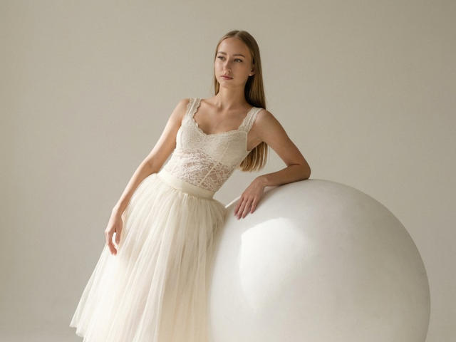 DaciaSchonhardt with long blonde hair, wearing a white lace dress and flowing skirt, leaning on a white sphere, showing her small tits and slim, attractive figure.