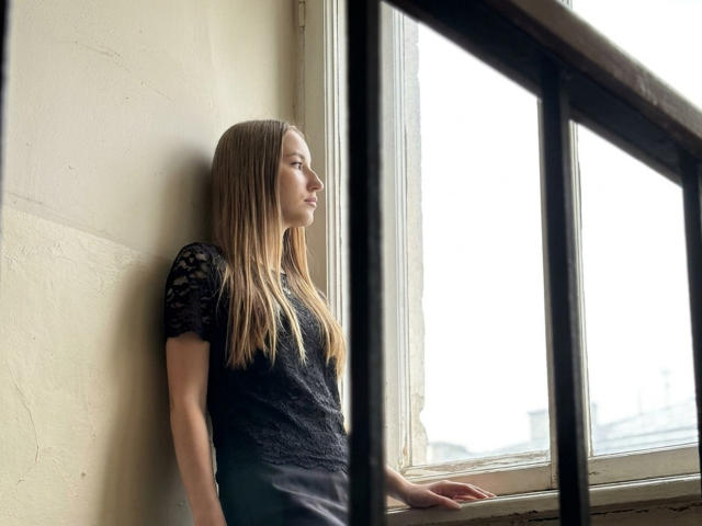 DaciaSchonhardt with long blonde hair, wearing a black lace top, standing by a window, looking outside, her hand on the sill, in a softly lit room.