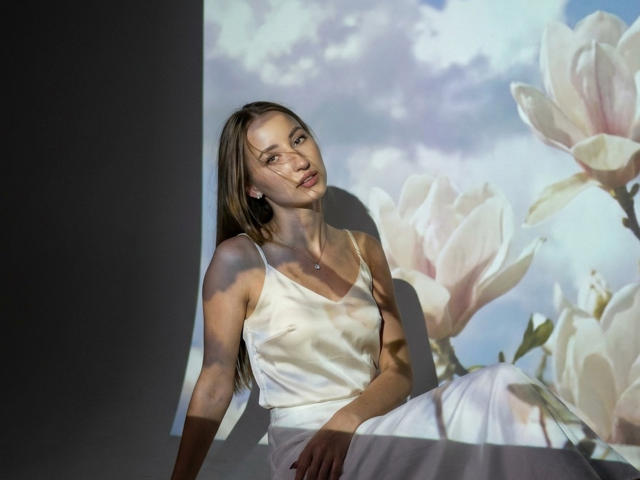 DaciaSchonhardt with long brown hair, wearing a white satin tank top and skirt, sitting, shadows on her face, soft light, flowers in background, relaxed, subtle smile.
