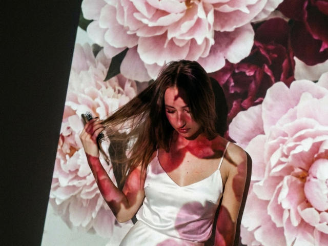 DaciaSchonhardt with long brown hair, holding it, wearing a white satin dress, standing in front of pink floral wallpaper, light casting pink shadows on her face and chest.