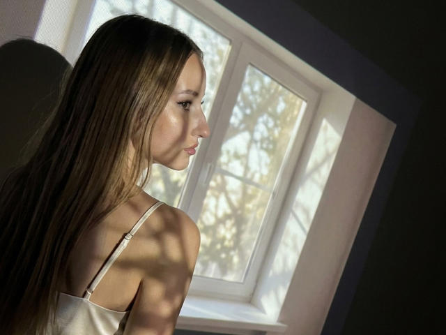 DaciaSchonhardt with long brown hair, white strap, looking out window, sunlight casting shadows on her face, upper back, and bare shoulders, in a quiet, dimly lit room.