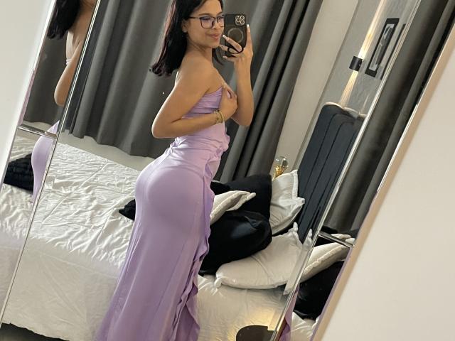 ChloeVixen in a lilac dress, holding her tits with one hand, showing her ass in a mirror selfie, wearing glasses, black hair, in a bedroom.
