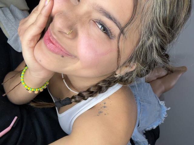 ChloeVixen with braided brown hair, smiling, nose ring, white tank top, yellow bead bracelet, hand on cheek, small tattoos on shoulder, pink lips, white necklace, denim sleeve.
