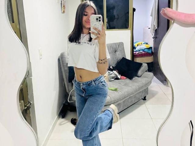 ChloeVixen with straight black hair, white crop top, blue jeans, and white socks, taking a mirror selfie, showing her flat stomach and tattoos, with one leg raised.